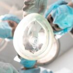 Three medical professionals in surgical masks and gowns, one of whom is holding an anesthetic inhaler close to the camera.