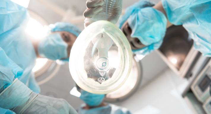 Three medical professionals in surgical masks and gowns, one of whom is holding an anesthetic inhaler close to the camera.