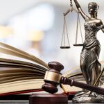 An open book and a gavel sit on a table next to a small statue of Lady Justice, blindfolded and holding a pair of scales.