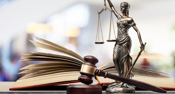 An open book and a gavel sit on a table next to a small statue of Lady Justice, blindfolded and holding a pair of scales.