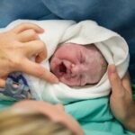 A blonde woman with an IV in her hand holding a newborn baby. The baby is crying, wrapped in a white blanket.