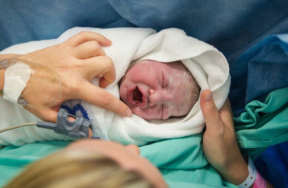 A blonde woman with an IV in her hand holding a newborn baby. The baby is crying, wrapped in a white blanket.