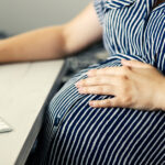 A visibly pregnant employee in a striped dress sitting at a computer desk, with one hand resting on her belly.
