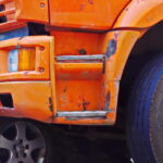 A close-up of an orange truck that has rear-ended a blue passenger car. The back end of the blue car is completely crumpled.