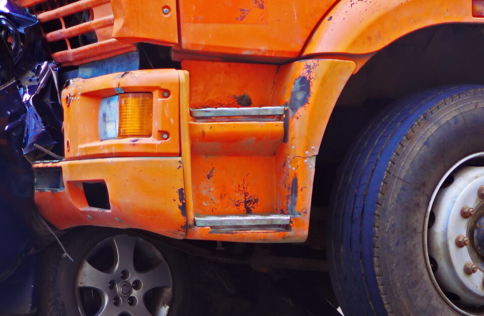 A close-up of an orange truck that has rear-ended a blue passenger car. The back end of the blue car is completely crumpled.