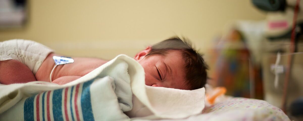 A newborn baby with dark hair lying on their stomach with their head turned to the side in a neonatal intensive care bed.