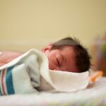 A newborn baby with dark hair lying on their stomach with their head turned to the side in a neonatal intensive care bed.