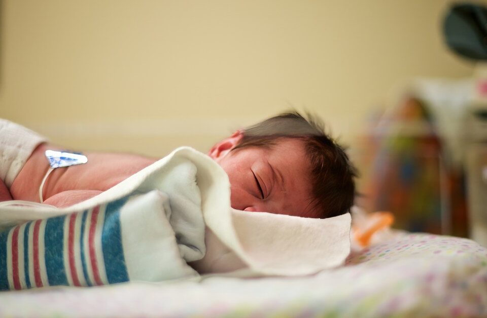 A newborn baby with dark hair lying on their stomach with their head turned to the side in a neonatal intensive care bed.