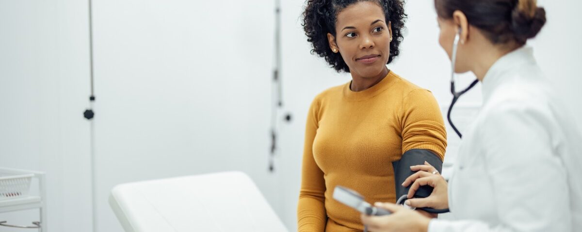 A white doctor in a lab coat measures the blood pressure of a young Black woman in an orange sweater.