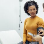 A white doctor in a lab coat measures the blood pressure of a young Black woman in an orange sweater.