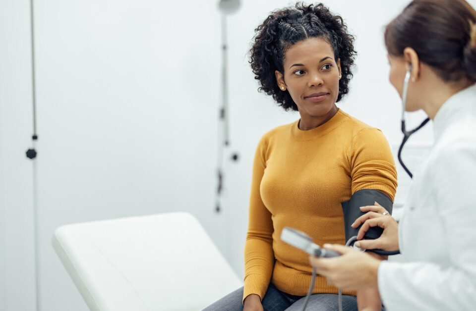 A white doctor in a lab coat measures the blood pressure of a young Black woman in an orange sweater.