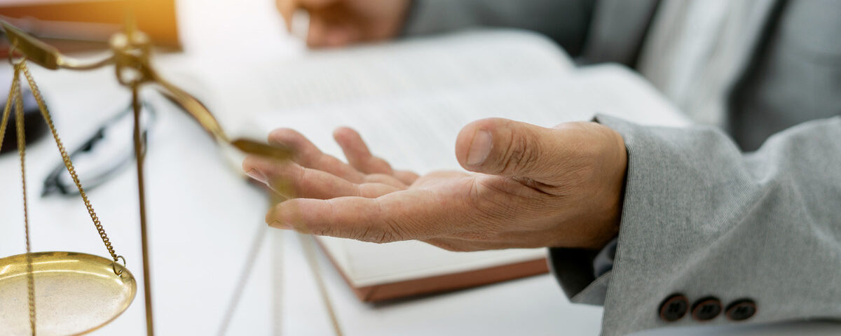 A person sits at a desk, with only their hands visible as they gesture with a pen. A small set of gold scales stands nearby.
