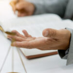 A person sits at a desk, with only their hands visible as they gesture with a pen. A small set of gold scales stands nearby.