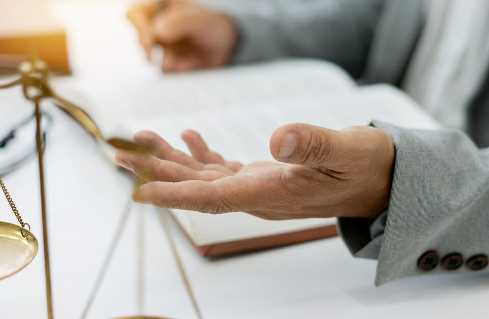 A person sits at a desk, with only their hands visible as they gesture with a pen. A small set of gold scales stands nearby.