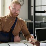 A young man with his right arm in a sling. He is sitting across a table from an attorney who has a laptop nearby.