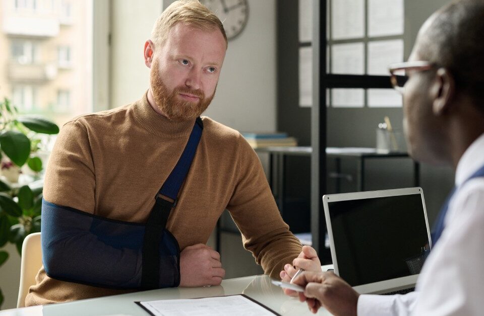 A young man with his right arm in a sling. He is sitting across a table from an attorney who has a laptop nearby.