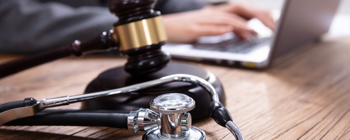 A gavel and a stethoscope arranged next to each other on a desk. Nearby, a person in a suit types on a laptop.