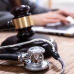 A gavel and a stethoscope arranged next to each other on a desk. Nearby, a person in a suit types on a laptop.