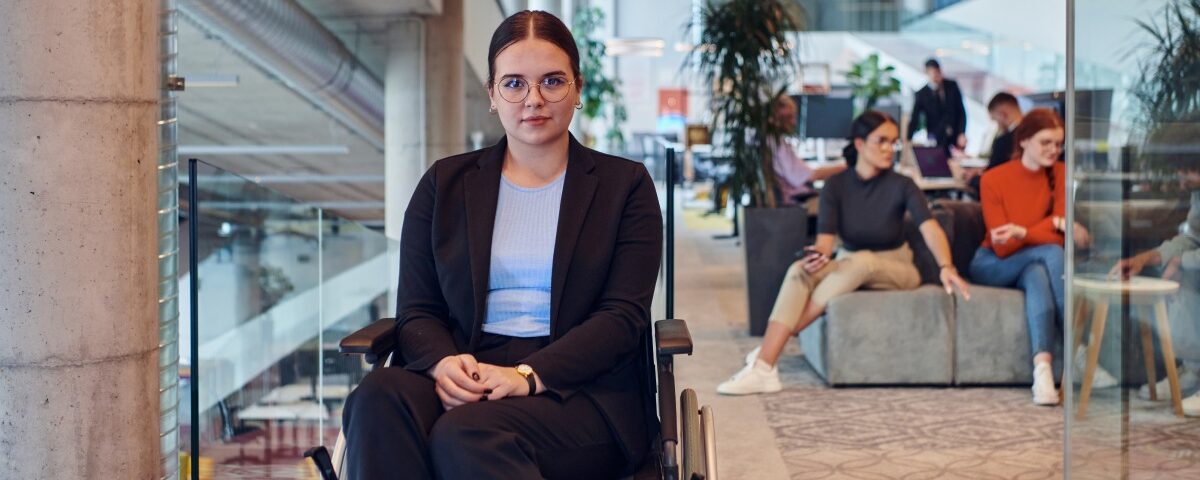 A woman with eyeglasses wears a suit and sits in her wheelchair, surrounded by other people in an open office space.