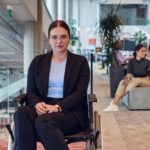 A woman with eyeglasses wears a suit and sits in her wheelchair, surrounded by other people in an open office space.
