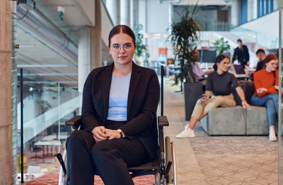 A woman with eyeglasses wears a suit and sits in her wheelchair, surrounded by other people in an open office space.