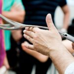A person with rolled-up sleeves holds out a pair of obstetrician’s forceps, showing three people what it looks like.