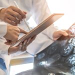 Two people in lab coats sitting at a desk. One is holding a digital tablet and the other is holding a scan of a human brain.