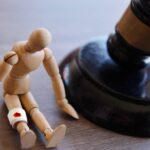 A gavel resting on a table next to a small wooden mannequin that is slumped over and has a gauze bandage around one leg.