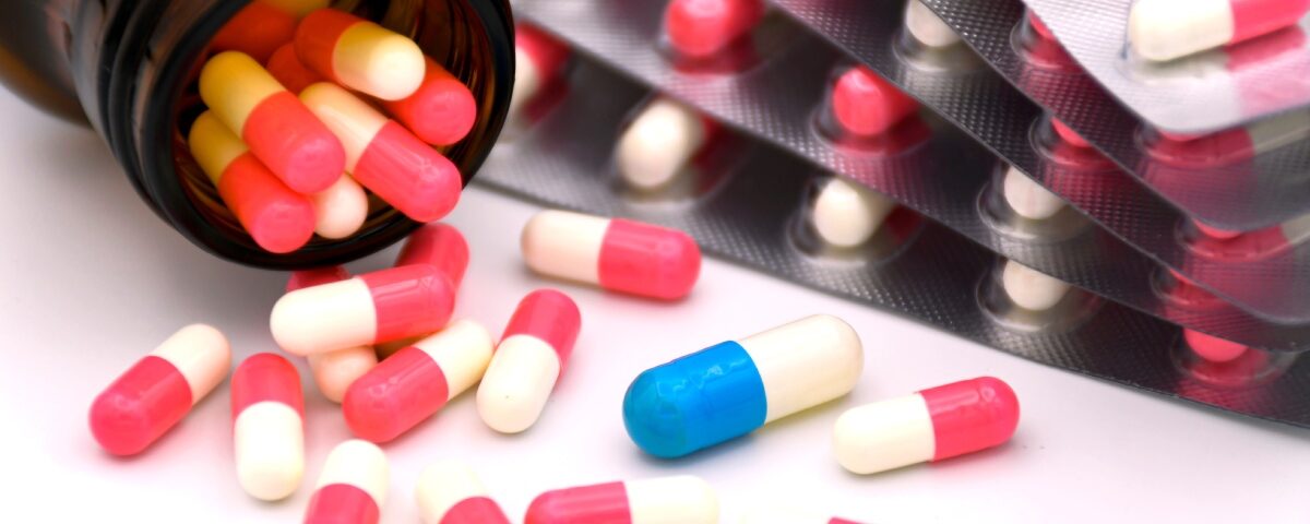 A medicine bottle tipped on its side with several capsules spilling onto a table. There is one blue capsule among many red ones.