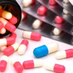 A medicine bottle tipped on its side with several capsules spilling onto a table. There is one blue capsule among many red ones.