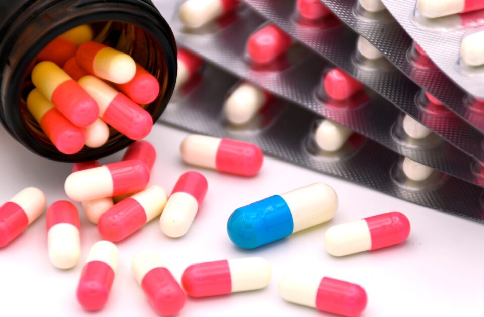 A medicine bottle tipped on its side with several capsules spilling onto a table. There is one blue capsule among many red ones.