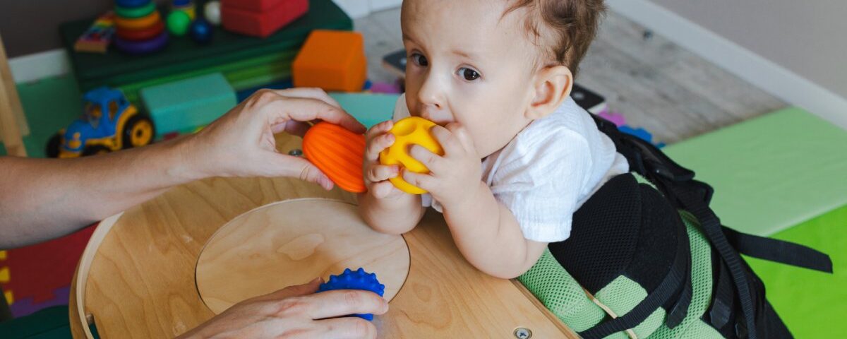 A baby steadying himself against a small table while gripping a yellow ball. An adult is holding other toys in front of him.