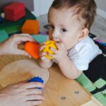 A baby steadying himself against a small table while gripping a yellow ball. An adult is holding other toys in front of him.