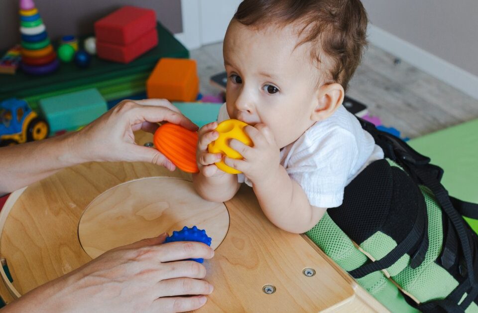 A baby steadying himself against a small table while gripping a yellow ball. An adult is holding other toys in front of him.
