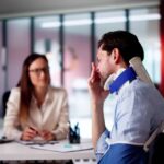 A man in a neck brace and an arm sling frowning as he speaks to a woman at a desk. She is listening intently to him.