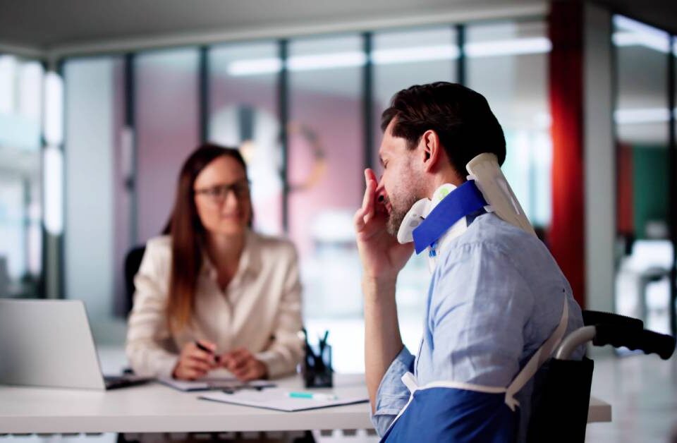 A man in a neck brace and an arm sling frowning as he speaks to a woman at a desk. She is listening intently to him.