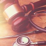 A stethoscope and a wooden gavel placed close together on a table. They are gently lit from the left by natural light.
