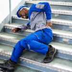 A man in blue work clothes and a hard hat lying on a metal staircase. He is wincing in pain as he grabs his lower back.
