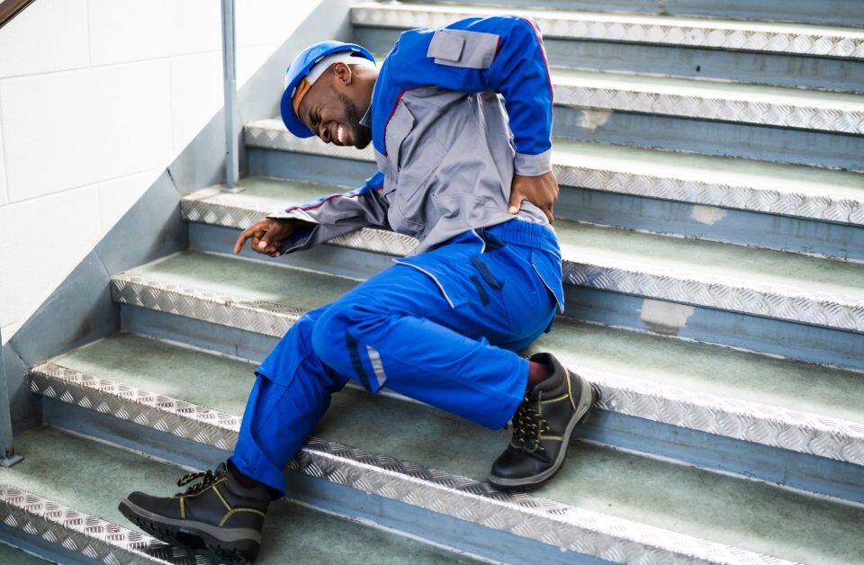 A man in blue work clothes and a hard hat lying on a metal staircase. He is wincing in pain as he grabs his lower back.