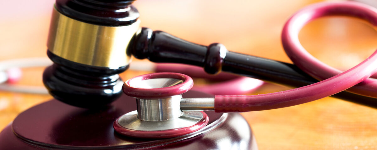A gavel rests on a sounding block on a wooden table. A red stethoscope is coiled around it and rests nearby.