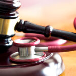 A gavel rests on a sounding block on a wooden table. A red stethoscope is coiled around it and rests nearby.