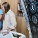 A person holds a brain scan of a patient. In the background, the patient, an elderly woman, is attended by a nurse.