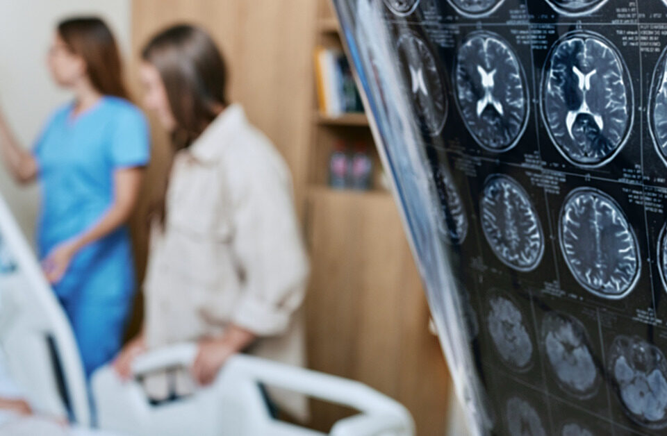 A person holds a brain scan of a patient. In the background, the patient, an elderly woman, is attended by a nurse.