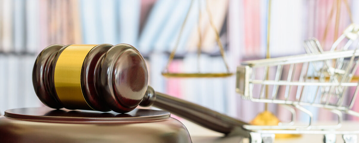 A gavel resting on a laptop next to a small set of gold scales and a miniature shopping cart. The setup is in a well-lit office.