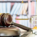A gavel resting on a laptop next to a small set of gold scales and a miniature shopping cart. The setup is in a well-lit office.