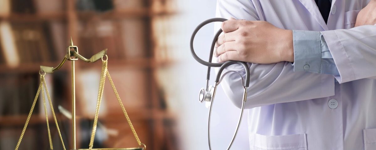 A person in a lab coat holds a stethoscope and crosses their arms. A small golden scale stands nearby.