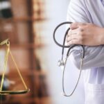 A person in a lab coat holds a stethoscope and crosses their arms. A small golden scale stands nearby.