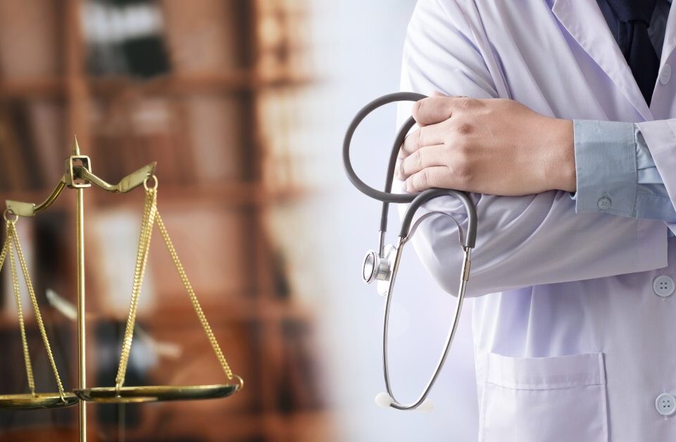 A person in a lab coat holds a stethoscope and crosses their arms. A small golden scale stands nearby.