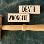 A plain wooden gavel lays horizontally on a marble table. Nearby are two wooden blocks that read “Wrongful” and “Death.”