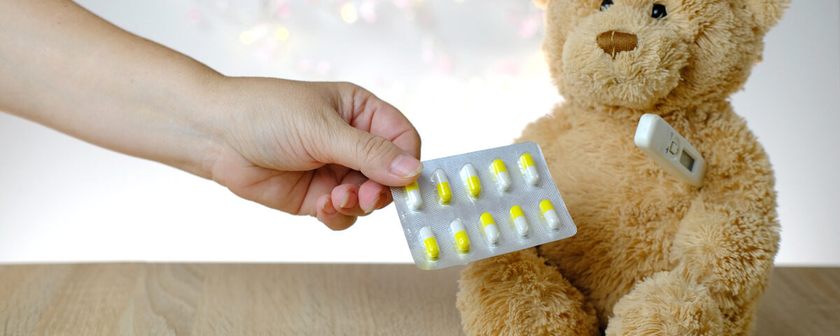 A teddy bear sits on a table with a thermometer under one arm, surrounded by loose pills and one blister pack of pills.